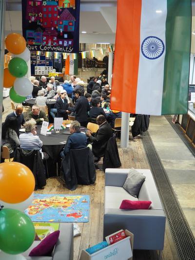 Groups of people gathered inside All Souls' church around tables, The National Flag of India hangs in the top right and childrens play corner can be seen in front of the groups of people.